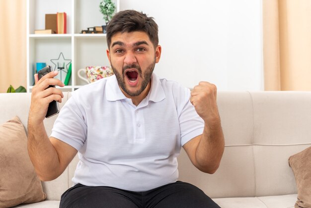 Young man in casual clothes holding smartphone clenching fist crazy happy and excited rejoicing his success sitting on a couch in light living room