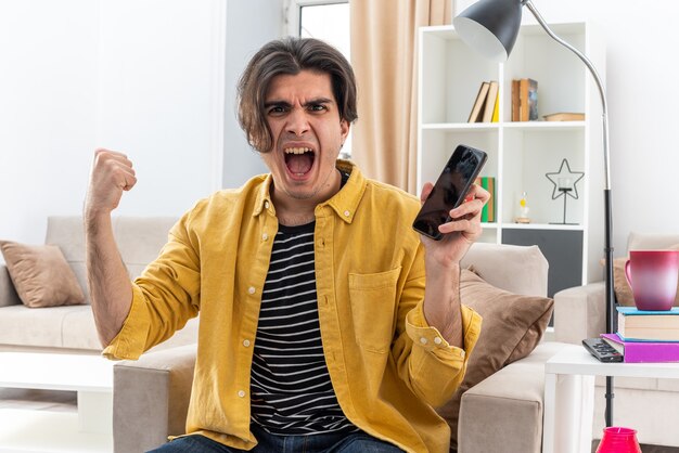 Young man in casual clothes holding smartphone clenching fist crazy mad shouting frustrated sitting on the chair in light living room