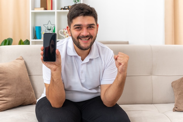 Young man in casual clothes holding smartphone clenching fist happy and excited rejoicing his success sitting on a couch in light living room