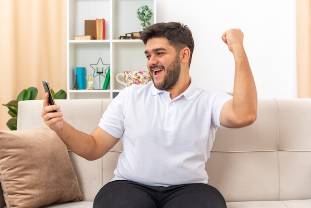 Young man in casual clothes holding smartphone clenching fist happy and excited sitting on a couch in light living room