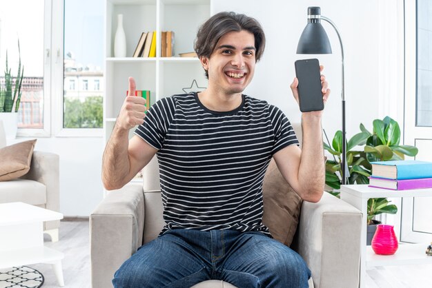 Young man in casual clothes holding smartphone  happy and cheerful showing thumbs up smiling broadly sitting on the chair in light living room