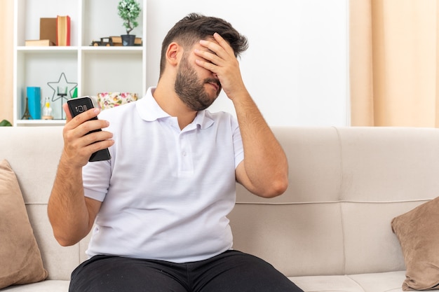 Young man in casual clothes holding smartphone looking annoyed and tired covering face with palm sitting on a couch in light living room