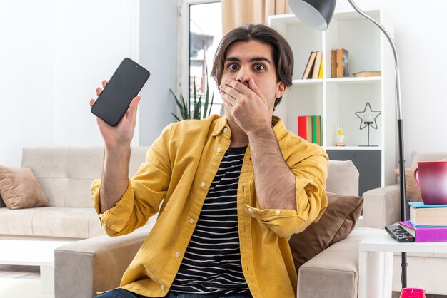 Young man in casual clothes holding smartphone looking being shocked covering mouth with hand sitting on the chair in light living room
