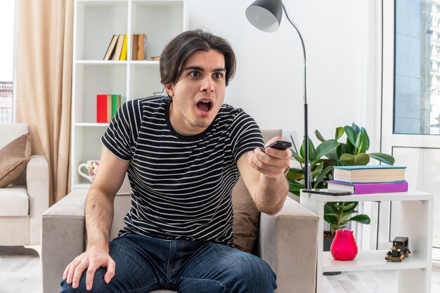 Young man in casual clothes holding tv remote looking amazed and surprised sitting on the chair in light living room