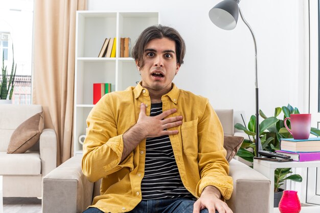 Young man in casual clothes looking amazed and surprised sitting on the chair in light living room