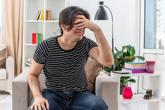 Free photo young man in casual clothes looking annoyed and irritated covering eyes with hand sitting on the chair in light living room