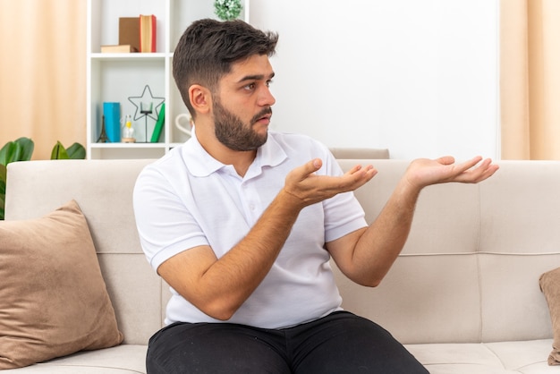 Young man in casual clothes looking aside confused presenting with arms something sitting on a couch in light living room