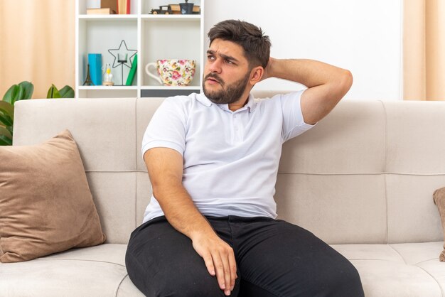 Young man in casual clothes looking aside confused with hand on his head sitting on a couch in light living room