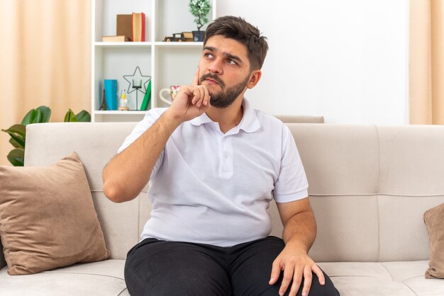 Young man in casual clothes looking aside puzzled sitting on a couch in light living room
