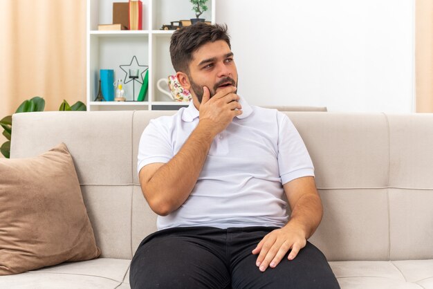 Young man in casual clothes looking aside with hand on chin puzzled sitting on a couch in light living room