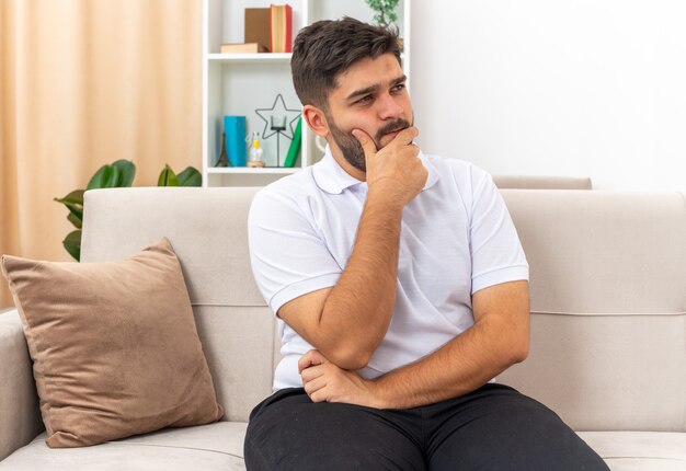 Young man in casual clothes looking aside with pensive expression with hand on chin thinking sitting on a couch in light living room