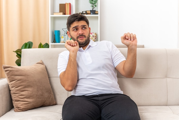 Free photo young man in casual clothes looking aside worried making defense gesture sitting on a couch in light living room