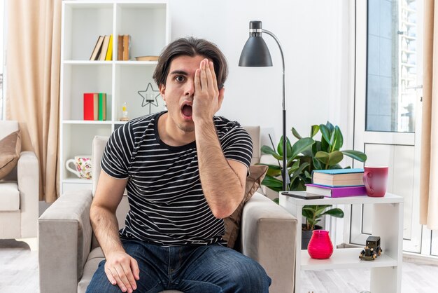 Young man in casual clothes looking confused covering one eye with hand sitting on the chair in light living room