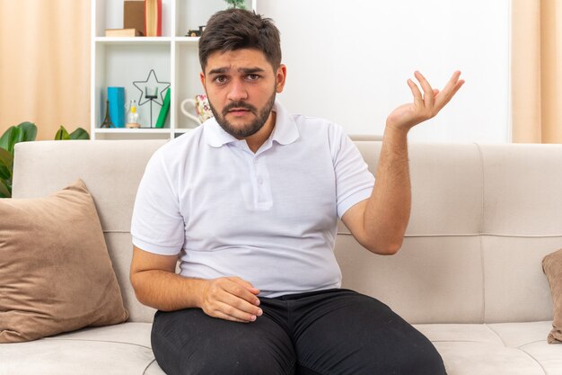 Young man in casual clothes looking  confused raising arm in displeasure and indignation sitting on a couch in light living room