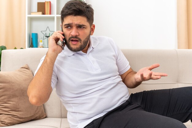 Young man in casual clothes looking confused while talking on mobile phone sitting on a couch in light living room