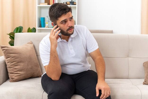 Young man in casual clothes looking confused while talking on mobile phone sitting on a couch in light living room