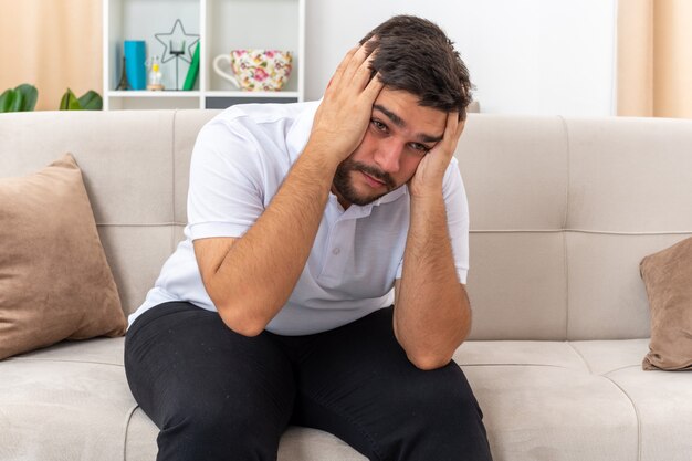 Young man in casual clothes looking depressed with hands on his head sitting on a couch in light living room