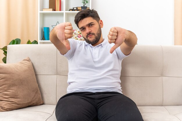 Young man in casual clothes looking displeased showing thumbs down sitting on a couch in light living room