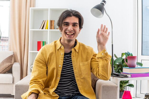 Young man in casual clothes looking happy and cheerful smiling broadly waving with hand sitting on the chair in light living room