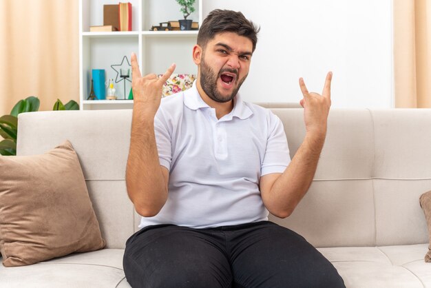 Young man in casual clothes looking happy and excited making rock symbol with fingers sitting on a couch in light living room