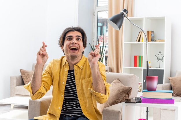 Young man in casual clothes looking happy and excited making wish crossing fingers sitting on the chair in light living room