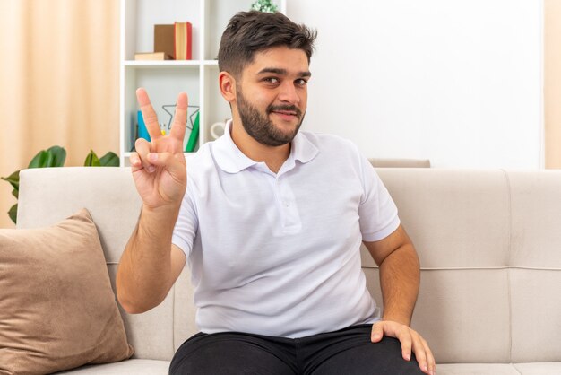 Young man in casual clothes looking  smiling cheerfully showing v-sign sitting on a couch in light living room