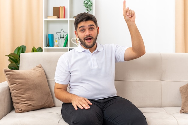 Молодой человек в повседневной одежде выглядит удивленным и счастливым, показывая указательный палец, имеющий новую идею, сидя на диване в светлой гостиной