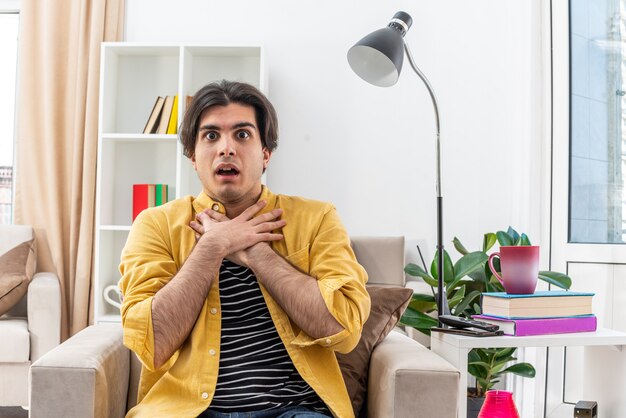 Young man in casual clothes looking surprised holding hands on his neck sitting on the chair in light living room