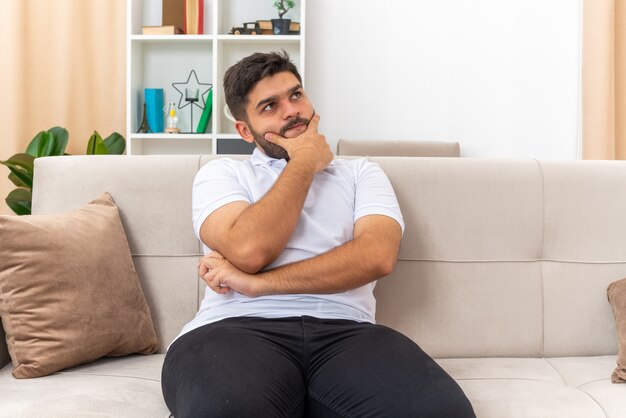 Young man in casual clothes looking up puzzled with hand on chin sitting on a couch in light living room