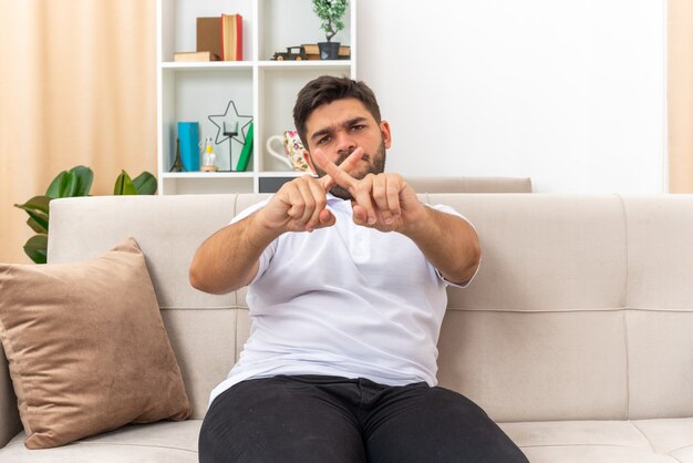 Young man in casual clothes looking with serious face making defense gesture crossing fingers sitting on a couch in light living room
