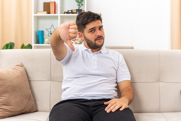 Young man in casual clothes looking  with serious face showing thumbs down sitting on a couch in light living room