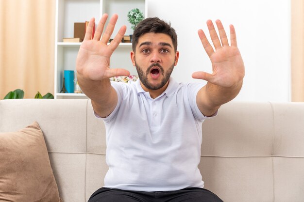 Young man in casual clothes looking worried and scared making stop gesture with hands sitting on a couch in light living room