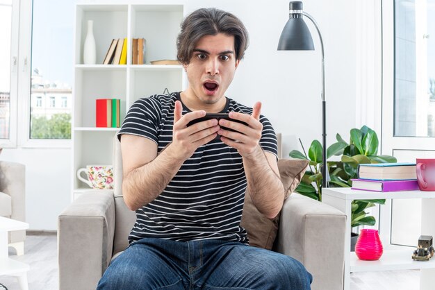 Young man in casual clothes playing games using smartphone amazed and excited sitting on the chair in light living room