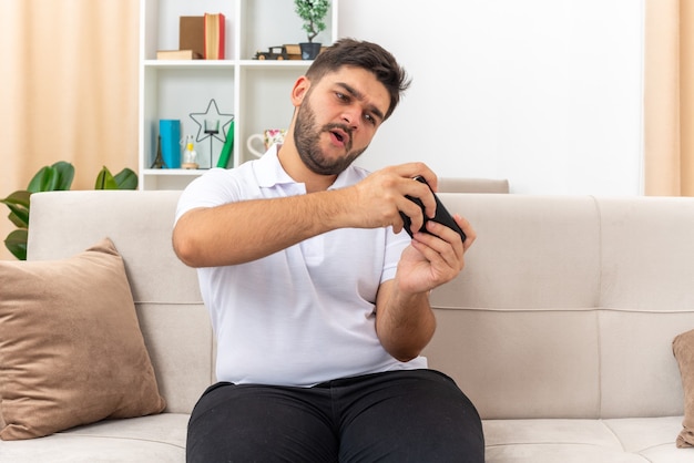 Молодой человек в повседневной одежде играет в игры с помощью смартфона, эмоционально и заинтригованно, сидя на диване в светлой гостиной