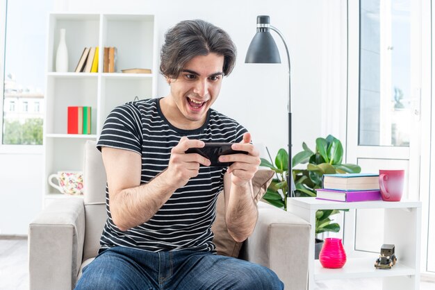 Young man in casual clothes playing games using smartphone happy and excited sitting on the chair in light living room