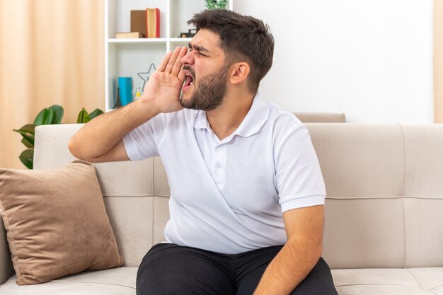 Young man in casual clothes shouting or calling with hand near mouth sitting on a couch in light living room