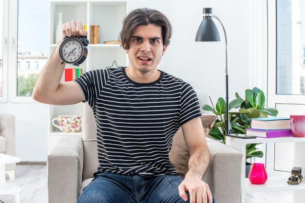 Young man in casual clothes showing alarm clock  confused and displeased sitting on the chair in light living room