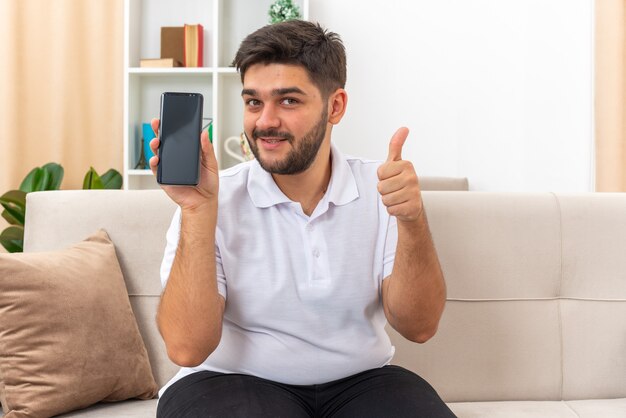 Молодой человек в повседневной одежде показывает смартфон, выглядит счастливым и позитивным, показывает палец вверх, сидя на диване в светлой гостиной