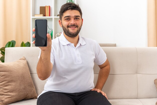 Молодой человек в повседневной одежде показывает смартфон, выглядящий счастливым и позитивным, улыбаясь, сидя на диване в светлой гостиной