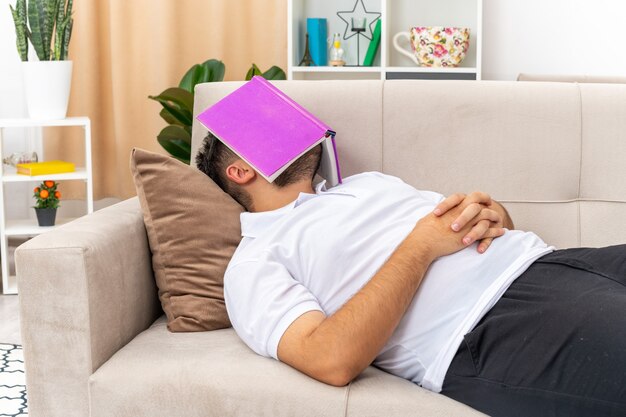 Young man in casual clothes with book over his head sleeping spending weekend at home laying on a couch in light living room