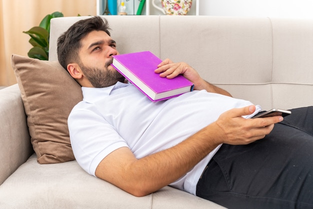 Young man in casual clothes with book looking up puzzled spending weekend at home laying on a couch in light living room