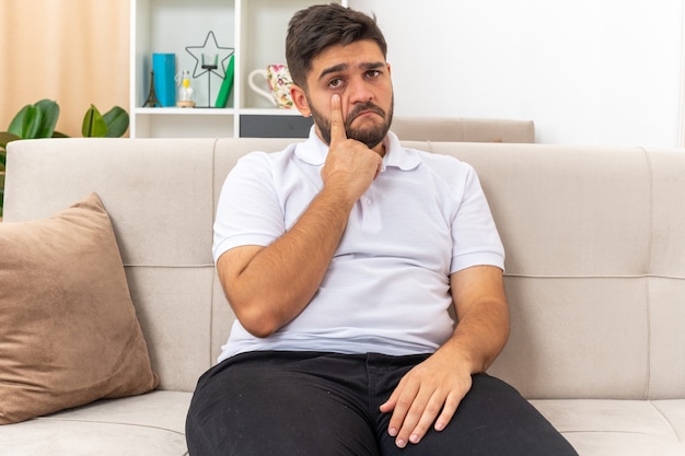 Young man in casual clothes  with sad expression pointing at his eye sitting on a couch in light living room