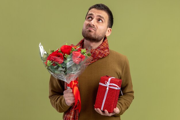 young man in casual clothes with scarf around neck holding bouquet of red roses and present looking up with sad expression valentines day concept standing over green wall