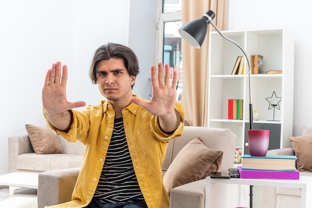 Young man in casual clothes  with serious face worried making stop gesture with hands sitting on the chair in light living room