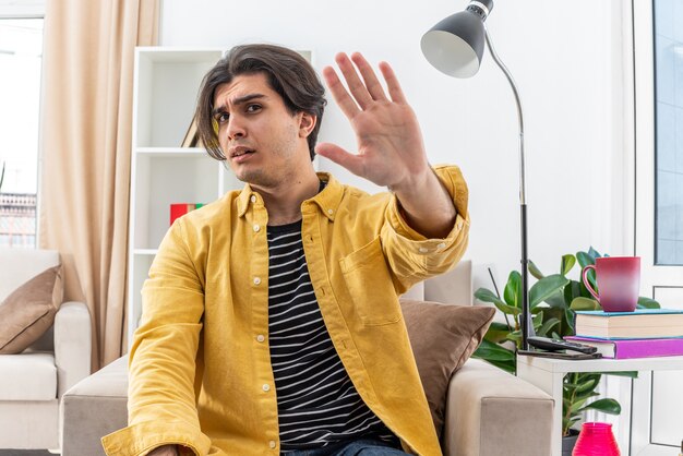 Young man in casual clothes  worried making stop gesture with hand sitting on the chair in light living room