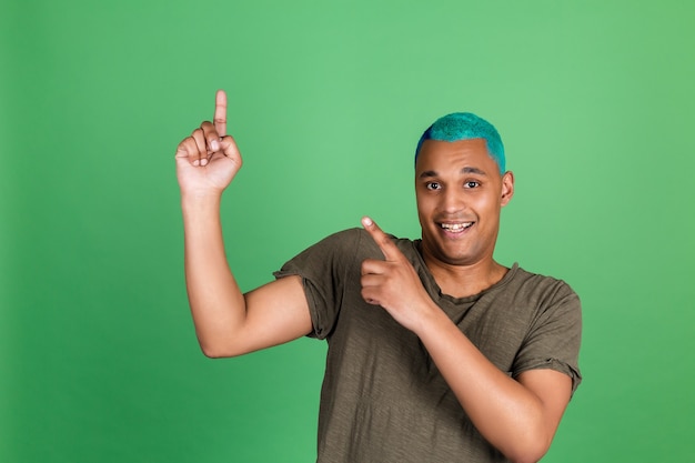 Молодой человек в повседневной одежде на зеленой стене с синими волосами указывает пальцем вверх