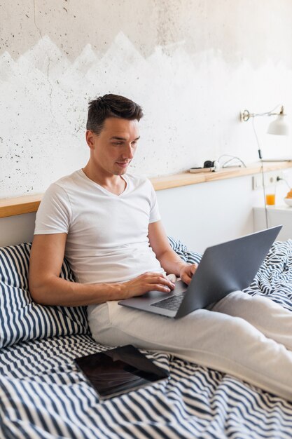 Young man in casual pajamas outfit sitting in bed in morning working on laptop, busy freelancer at home