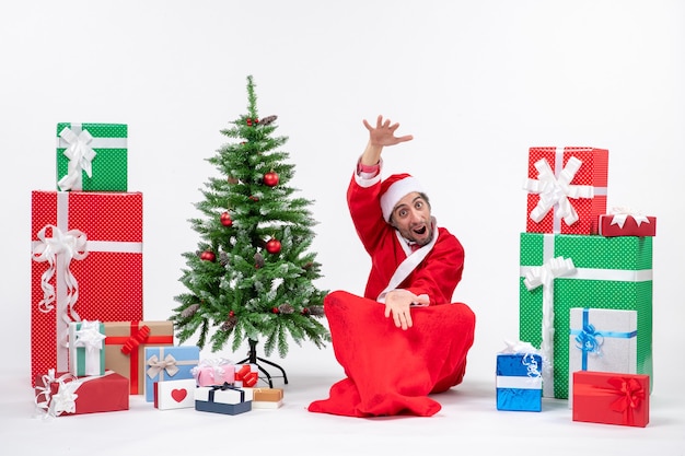 Молодой человек празднует рождество, сидя в земле, показывая что-то рядом с подарками и украшенной рождественской елкой