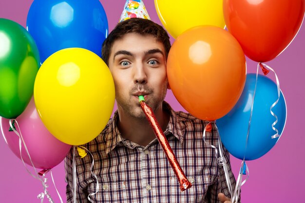 Young man celebrating birthday, holding colorful baloons over purple wall.