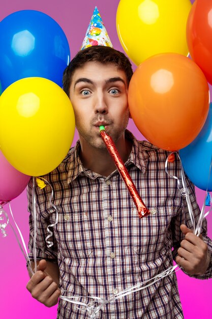 Young man celebrating birthday, holding colorful baloons over purple wall.